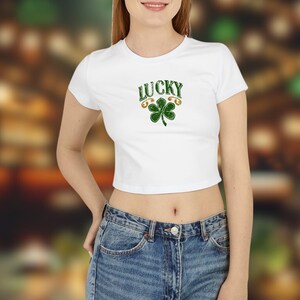 Könnte beinhalten: Weißes Crop-Top mit dem Wort "LUCKY" in Grün, über einem grünen Kleeblatt mit goldenen Akzenten. Das Shirt wird mit blauen Jeans kombiniert. Ein festliches Design für St. Patrick's Day.