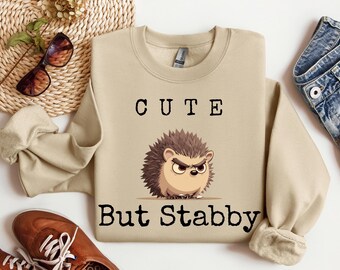 Könnte beinhalten: Ein beiger Pullover mit einem Comic-Igel und dem Text "Cute But Stabby".
