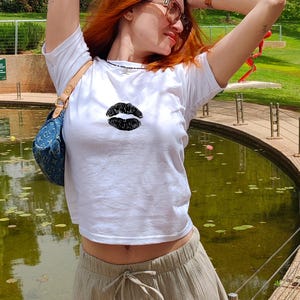 May include: A woman wearing a white cropped t-shirt with a black lip print design. She is wearing a light brown skirt and has her arms raised above her head. The background is a pond with lily pads and a green grassy area.