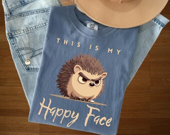 Könnte beinhalten: Ein blaues T-Shirt mit dem Text "THIS IS MY Happy Face" und einer Illustration eines Igels. Das T-Shirt liegt auf einer blauen Jeans und einem beigen Hut.
