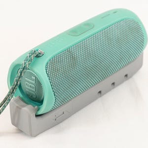 May include: A teal blue portable Bluetooth speaker with a gray base and a green and gray rope strap.