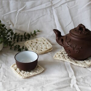 May include: A brown teapot with a floral design sits on a crocheted coaster. A small brown cup sits on a similar coaster. A potted plant with green leaves is in the background.