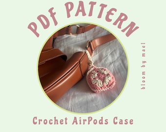 Op de afbeelding: Een gehaakt AirPods-hoesje met een roze en wit bloemenmotief. Het hoesje is bevestigd aan een bruine leren tas met een band. De afbeelding bevat de tekst "PDF PATTERN bloom by mael Crochet AirPods Case".