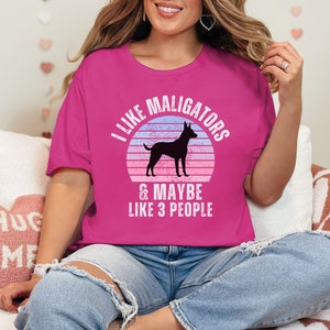 May include: Berry-colored t-shirt with the text "I LIKE MALIGATORS & MAYBE LIKE 3 PEOPLE" and a silhouette of a dog inside a sunset design. The shirt is a casual, relaxed fit.
