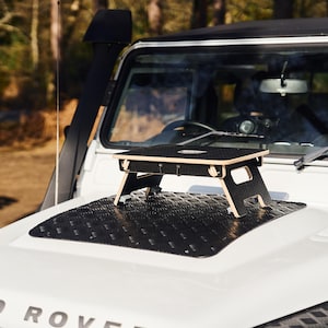 May include: A black folding table with a wooden top sits on the bonnet of a white Land Rover. The bonnet is covered in a black diamond-patterned mat.