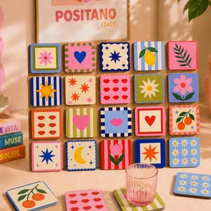 Può includere: Una collezione di sottobicchieri quadrati colorati con vari disegni, tra cui fiori, cuori e frutta. Sono visibili anche un bicchiere trasparente a righe e una pila di libri. I sottobicchieri sono disposti su una superficie chiara.