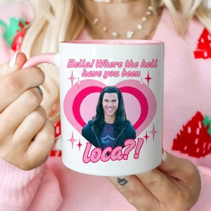 Puede incluir: Taza de cerámica blanca con interior y asa rosas. La taza presenta un gráfico de la cara de un hombre dentro de un corazón rosa, con el texto "Bella! Where the hell have you been" y "Loca?!" en rosa.