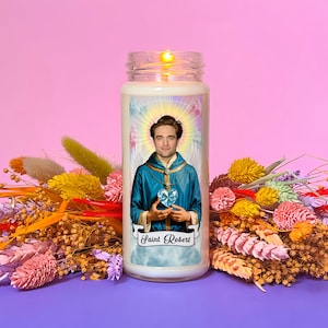 May include: A white candle with a picture of a man wearing a blue robe and holding a blue heart. The candle is lit and the text "Saint Robert" is printed on the front.