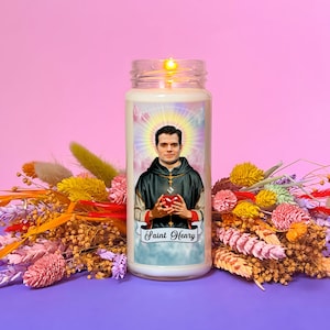 Puede incluir: Una vela blanca con una imagen de un hombre con una túnica oscura sosteniendo un corazón rojo. La vela tiene el texto "Saint Henry" en la parte delantera. La vela está rodeada de flores secas.