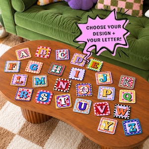 Pode incluir: Uma coleção de vinte e seis porta-copos quadrados, cada um apresentando uma letra única do alfabeto de A a Z em um padrão colorido distinto. Um gráfico de balão de fala rosa com o texto preto CHOOSE YOUR DESIGN + YOUR LETTER! está incluído.