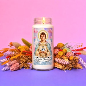 May include: A lit, white pillar candle with a label featuring a portrait of a man with a halo, wings, and a blue cape, holding a globe. The text "Saint Connor" is below the image. The candle is surrounded by colourful dried flowers.