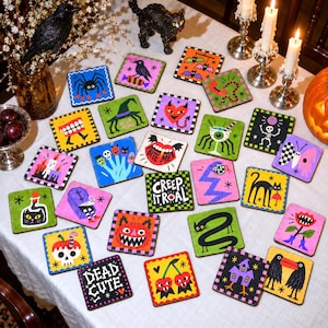Może przedstawiać: Kolekcja kwadratowych podkładek z kolorowymi ilustracjami motywów halloweenowych, w tym pająków, czaszek, nietoperzy, czarnych kotów, duchów oraz napisów takich jak BITE ME, CREEP IT REAL i DEAD CUTE na białym obrusie.