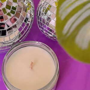 Może przedstawiać: Mała, biała świeca w przezroczystym szklanym słoiku z drewnianym knotem. Świeca jest otoczona dwiema srebrnymi kulami dyskotekowymi i zielonym liściem.