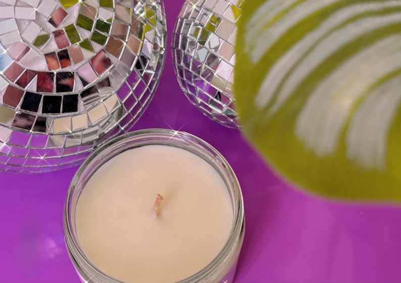 Może przedstawiać: Mała, biała świeca w przezroczystym szklanym słoiku z drewnianym knotem. Świeca jest otoczona dwiema srebrnymi kulami dyskotekowymi i zielonym liściem.