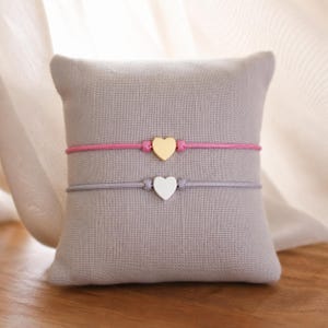 Peut inclure: Deux bracelets en cordon avec des breloques en forme de cœur. Le bracelet du haut possède un cordon rose vif avec un cœur doré, tandis que celui du bas a un cordon gris clair avec un cœur argenté. Les deux sont centrés sur un coussin carré.
