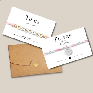 Peut inclure: Deux cartes de vœux avec des bracelets et une enveloppe marron. L'une dit "Tu es une cousine en or" avec un bracelet rose. L'autre dit "Tu vas être" avec un bracelet à motifs et un charm argenté.