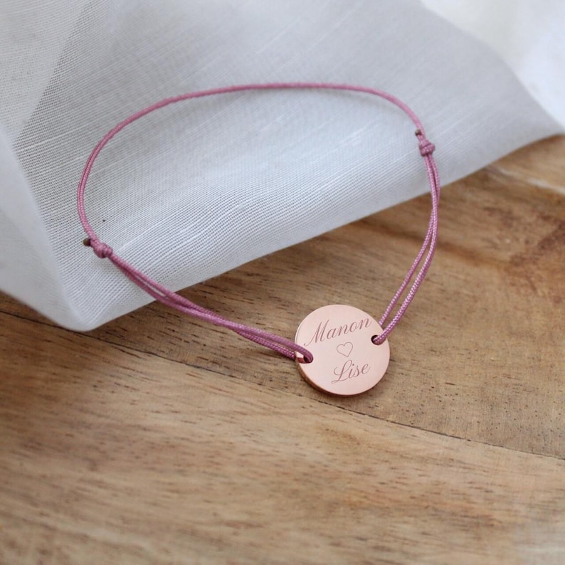 May include: A pink string bracelet with a round, rose gold charm engraved with the text "Manon &hearts; Lise".