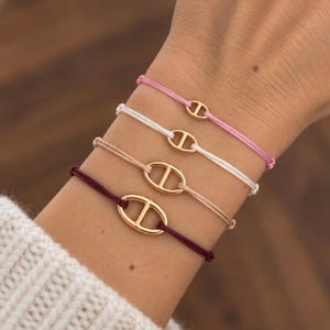 Peut inclure: Quatre bracelets en cordon dotés de maillons métalliques dorés de forme ovale avec une barre horizontale centrale. Les cordons sont dans des tons rose clair, blanc, beige et bordeaux foncé, chacun fixé par un mécanisme de nœud coulissant.