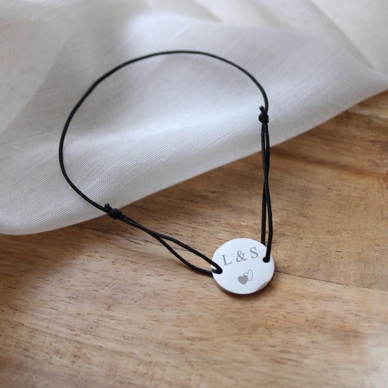 May include: A black cord bracelet with a silver round charm engraved with the initials "L & S" and two small hearts.
