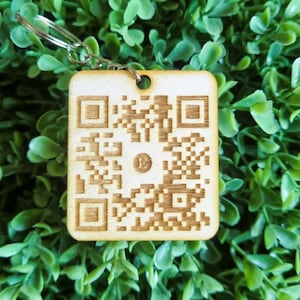 Peut inclure: Un porte-clés en bois avec un code QR gravé au laser. Le porte-clés est brun clair et possède un anneau en métal argenté.