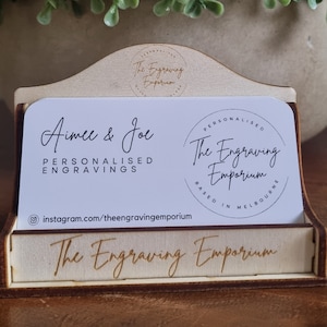 May include: A wooden business card holder with a light brown finish. The holder displays business cards with the text "Aimee & Joe" and "The Engraving Emporium." The holder is on a wooden surface, with a green plant in the background.
