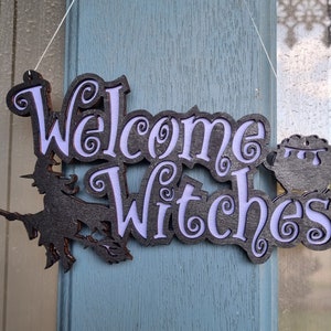 Puede incluir: Un letrero de madera negro con las palabras "Welcome Witches" en letras moradas. Una silueta de bruja vuela en una escoba a la izquierda del letrero. Un caldero está a la derecha del letrero.