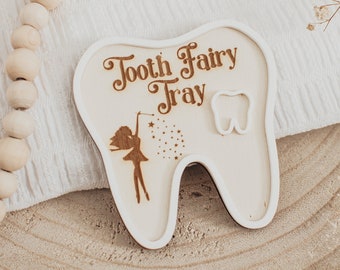 May include: Wooden tooth fairy tray in the shape of a tooth. The tray has the words "Tooth Fairy Tray" engraved on it, along with an illustration of a fairy and a tooth. The tray is made of light-colored wood.