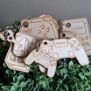 May include: Five wooden keychains shaped like video game controllers and a game console. The keychains are engraved with the names "Leo", "Daddy", "Dadda", "Jack", and "Ddy".