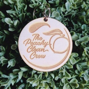 Op de afbeelding: Een ronde houten sleutelhanger met de tekst "The Peachy Clean Crew" en een perzikafbeelding. De sleutelhanger is lichtbruin en hangt aan een zilveren metalen ring. De achtergrond is een dichte verzameling groene bladeren.