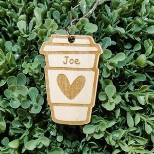 May include: Wooden coffee cup keyring with the name "Joe" engraved on it. The keyring has a heart shape cut out on the front.
