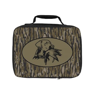 May include: A rectangular lunch bag with a black handle and zipper. The bag features a camouflage pattern in shades of brown and green. An oval emblem displays a black silhouette of a dog holding a duck in its mouth.