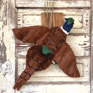 May include: A plush pheasant wall hanging with outstretched wings. The bird features brown, black, blue, green, and white coloring. It is mounted on a small wooden plaque with dried grass accents, perfect for rustic decor.