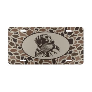 May include: License plate with a hunting dog illustration. The oval center features a dog holding a pheasant, in shades of brown and black. The background has a camouflage pattern in brown and tan.