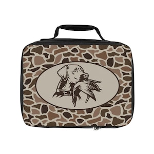 May include: A rectangular lunch bag with a black handle and zipper. The bag features a camouflage pattern in shades of brown and tan. An oval graphic depicts a dog holding a duck in its mouth.