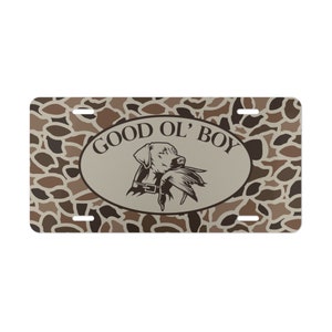 May include: A brown and tan camouflage patterned license plate with a white oval in the center. The oval has the text "GOOD OL' BOY" in brown text and a black and white illustration of a dog with a duck in its mouth.