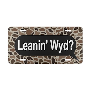 Op de afbeelding: Een bruin en tan camouflage-patroon kentekenplaat met een zwarte spraakballon die "Leanin' Wyd?" zegt in witte tekst.
