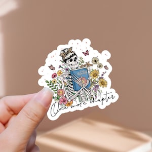 Può includere: Un adesivo con uno scheletro che tiene un libro, circondato da fiori e farfalle. Il testo "One more chapter" è scritto sotto lo scheletro.