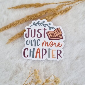 Könnte beinhalten: Weißer Aufkleber mit dem Text "Just one more chapter" in brauner, lila und grauer Schrift. Oberhalb des Textes befindet sich eine kleine Illustration eines geöffneten Buches mit einem Blattzweig.