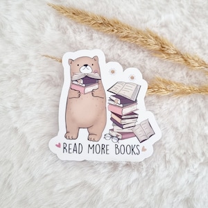 Könnte beinhalten: Ein brauner Bär mit einem Schnurrbart hält einen Stapel Bücher. Ein zweiter Stapel Bücher befindet sich neben dem Bären. Der Aufkleber lautet "Lies mehr Bücher".