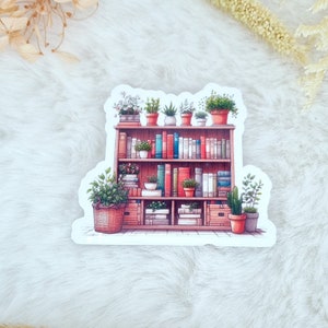 Könnte beinhalten: Ein Bücherregal aus Holz mit drei Regalbrettern, gefüllt mit Büchern und Topfpflanzen. Das Bücherregal ist hellbraun lackiert und hat einen weißen Hintergrund.