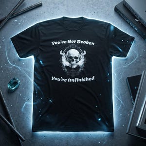 Pu&ograve; includere: Una t-shirt nera a maniche corte con una grafica bianca di un teschio centrato all'interno di una cornice ovale decorata. Il testo "You're Not Broken" appare sopra il teschio e "You're Unfinished" &egrave; stampato sotto in lettere bianche.