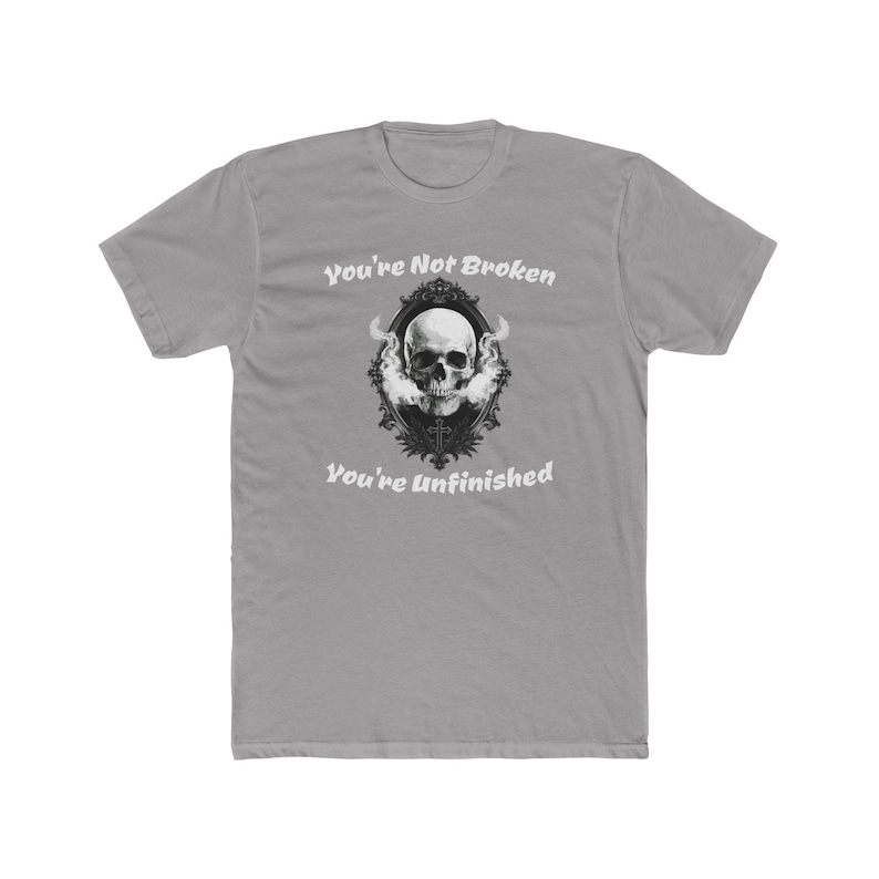 Pu&ograve; includere: Una t-shirt grigia a maniche corte con la grafica di un teschio umano all'interno di una cornice ovale nera decorata. Il teschio &egrave; circondato da fumo bianco. Il testo "You're Not Broken" appare sopra e "You're Unfinished" sotto.