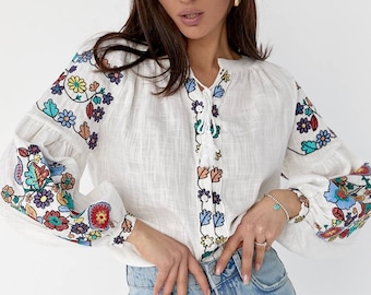 Puede incluir: Blusa blanca de lino con bordado floral de colores en las mangas y el escote. La blusa tiene cuello en V y un lazo en el escote.
