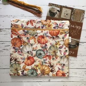 May include: A square fabric pouch featuring a repeating pattern of pumpkins, autumn leaves, and berries in shades of orange, green, and cream. The pouch has a gold-toned zip closure with a thin orange pull tab at the top edge of the bag.