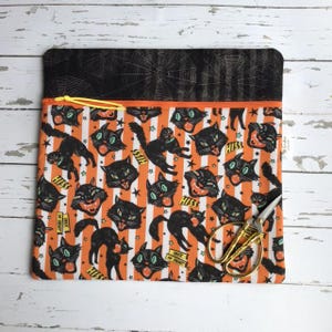 May include: A rectangular fabric pouch featuring a black top panel with a spiderweb pattern and a main body with orange and white vertical stripes. The print displays black cats, the text HISS, and TRICK OR TREAT. A yellow zipper is attached.