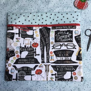 May include: A rectangular fabric pouch with a light blue top panel featuring black dots and a main body printed with vintage sewing-themed graphics, including sewing machines, top hats, eyeglasses, and text, secured by a bright red zipper.