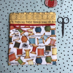 May include: A rectangular fabric pouch with a red zipper, featuring a white background with a colorful print of various sewing thread spools and text. The top border is yellow with a ruler pattern and the text B. THEN ALL THESE THINGS SHALL BE ADDED.
