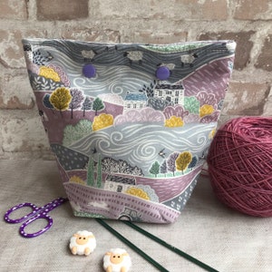 May include: A gray and purple fabric project bag with a whimsical countryside print featuring houses, trees, and sheep. The bag has purple buttons and is filled with yarn and knitting needles.