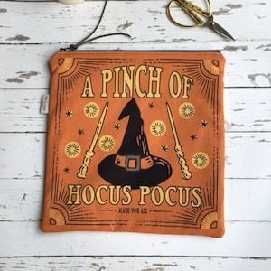 May include: A square orange zippered pouch featuring a black witch hat graphic in the centre, flanked by two wands. The text reads A PINCH OF HOCUS POCUS and MAGIC FOR ALL in a decorative frame with star and sunburst accents printed in black ink.