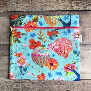 May include: A square zippered pouch featuring a light blue background with a colorful pattern of hot air balloons, various orange and pink flowers, green leaves, and blue birds. A bright pink zipper runs across the top with a yellow cord pull.